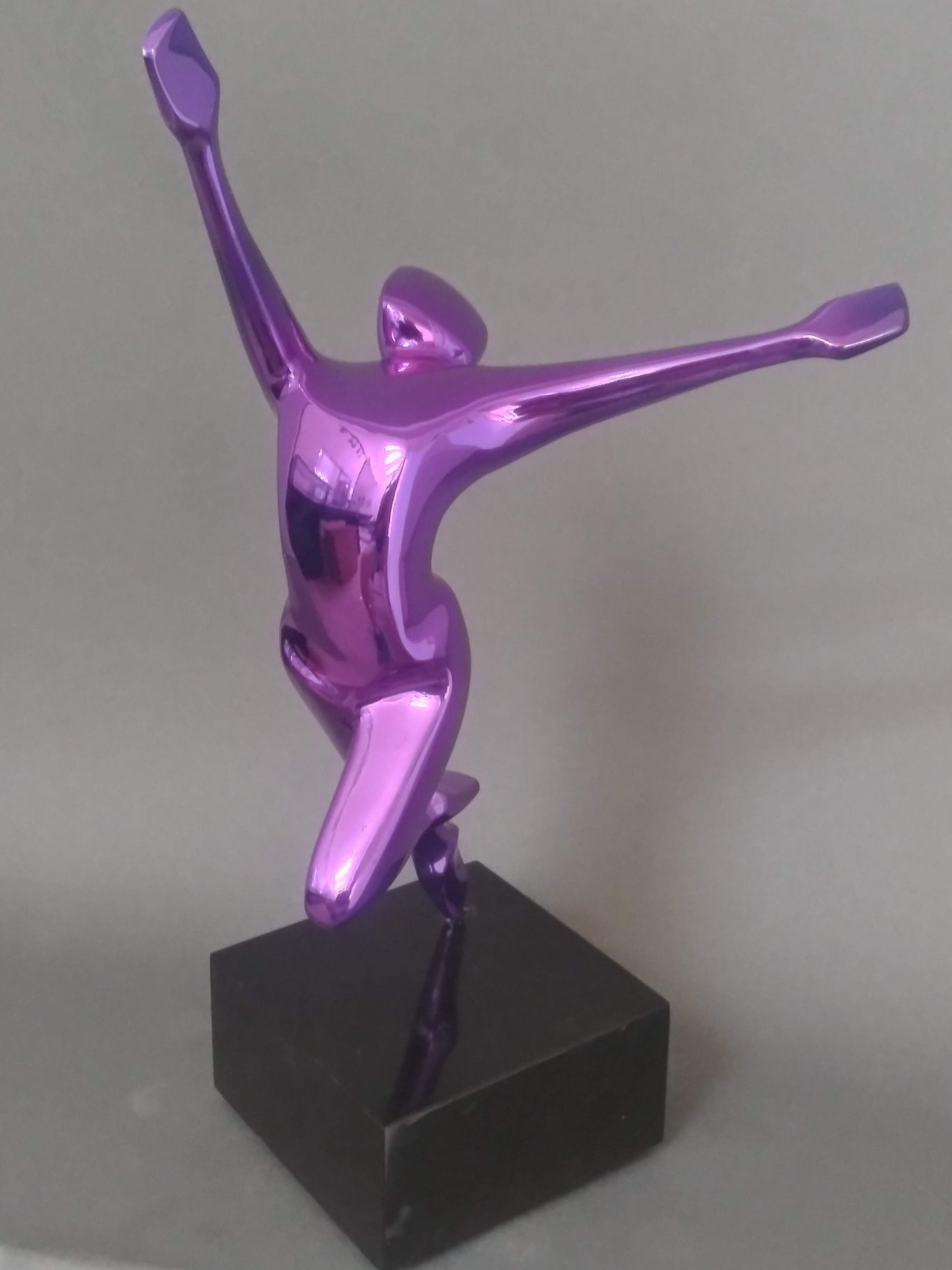 A purple metallic sculpture of an abstract human figure with arms outstretched in a joyful pose, mounted on a black marble base.