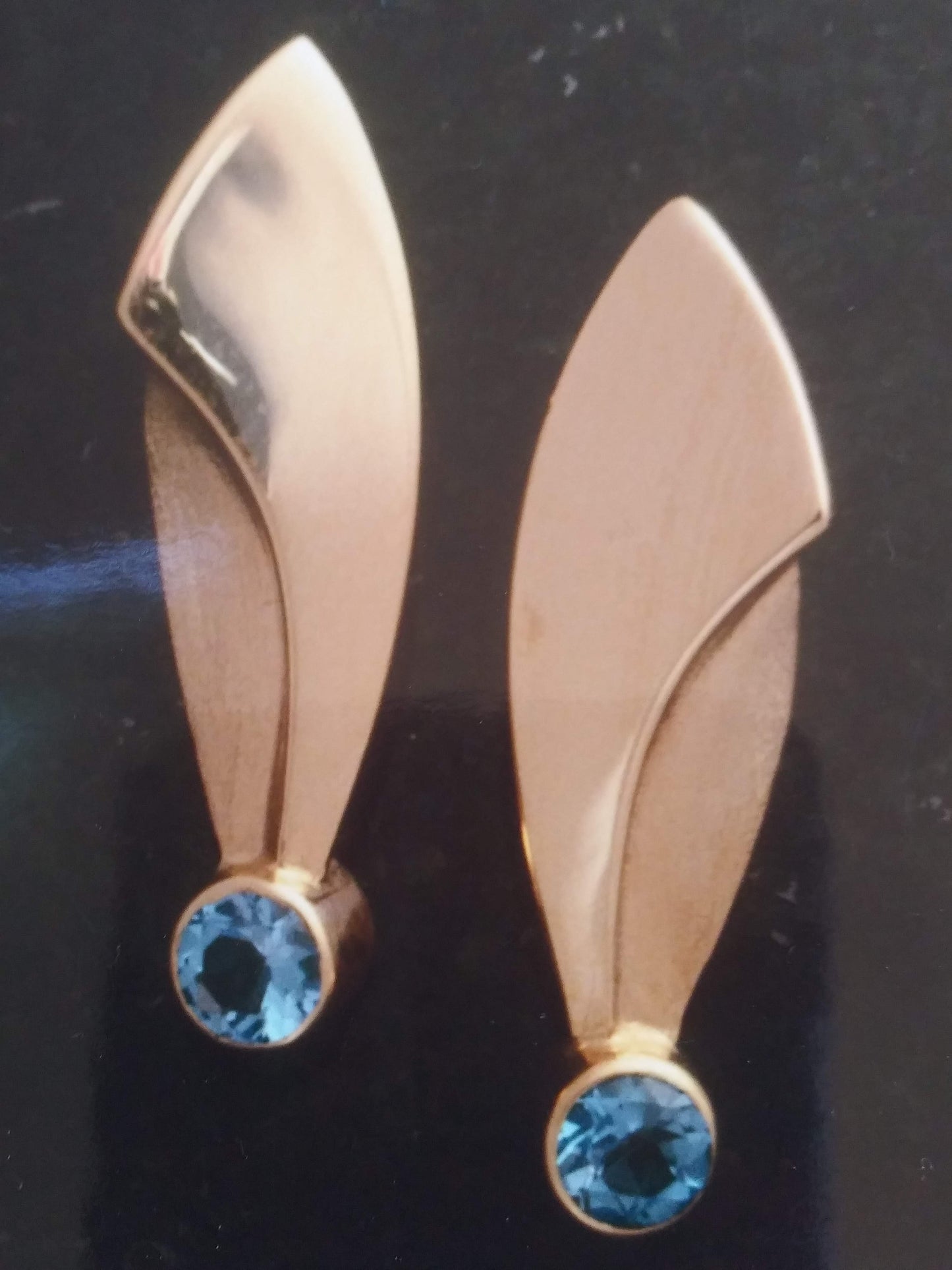 A pair of gold earrings with a curved, leaf-like design and a single blue gemstone at the bottom of each earring.