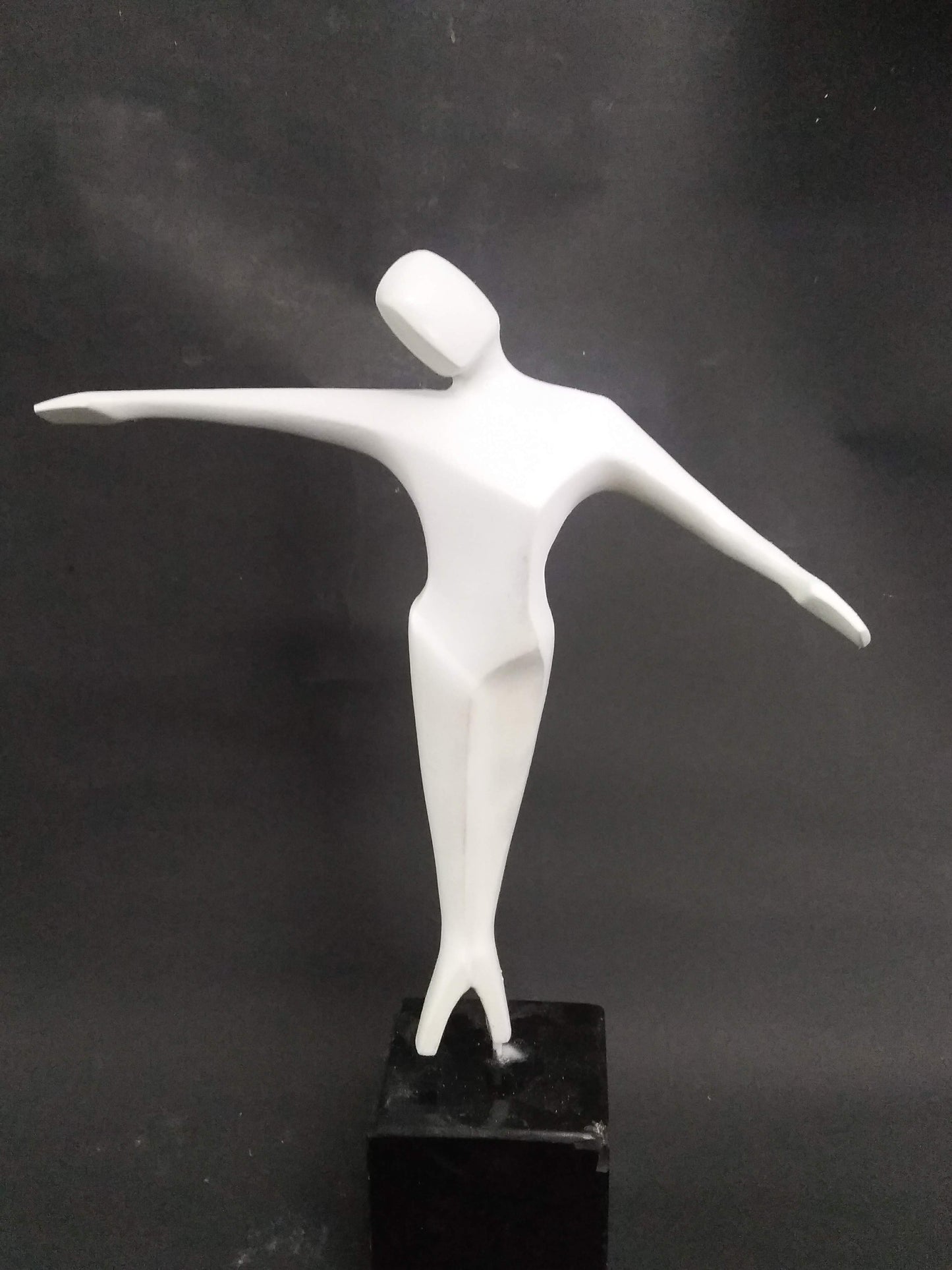 A white abstract sculpture of a human figure with arms outstretched, standing on a black pedestal.