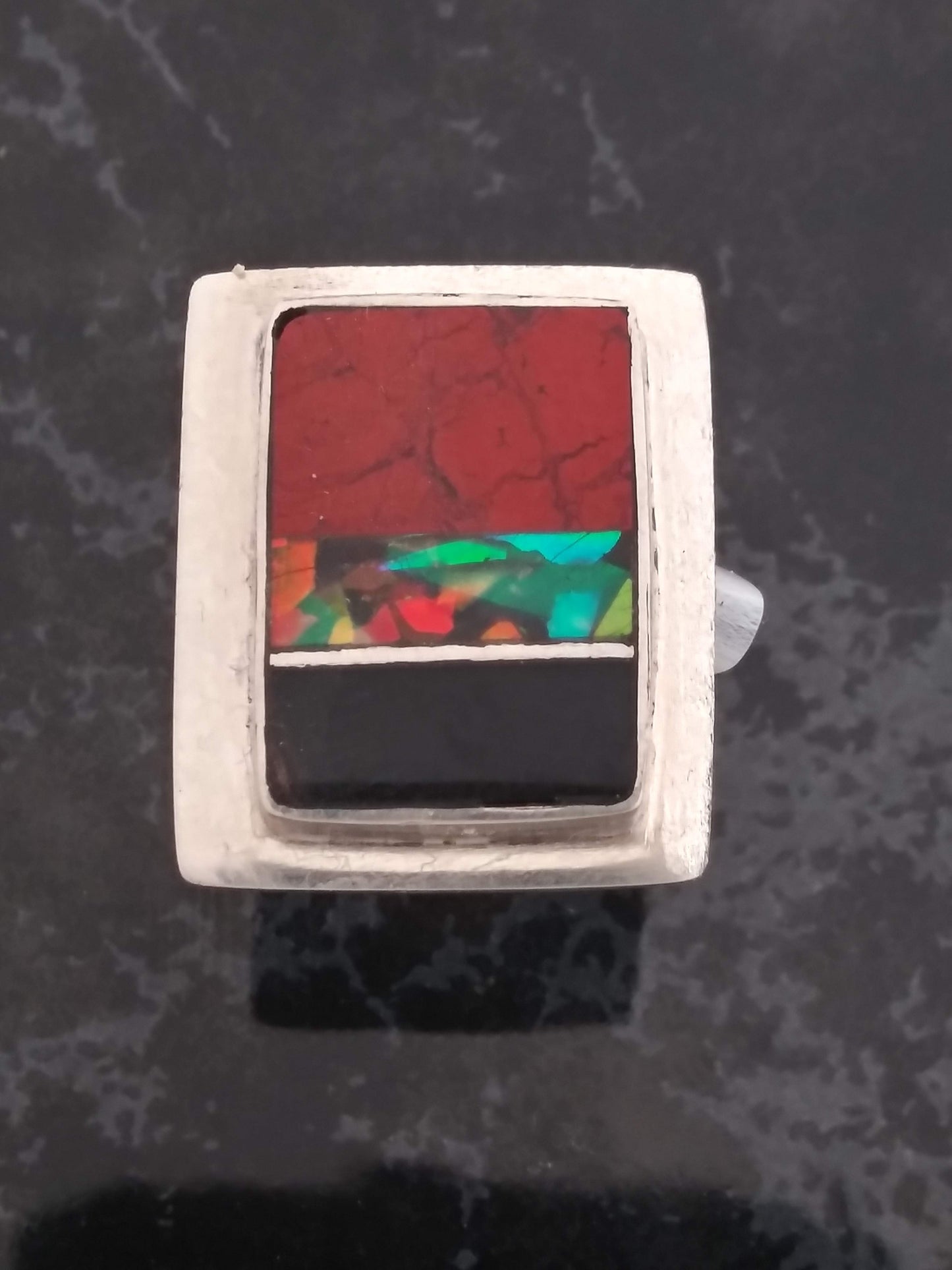 A silver ring with a rectangular inlay featuring a red stone at the top, a thin band of colorful opal in the center, and a black stone at the bottom.