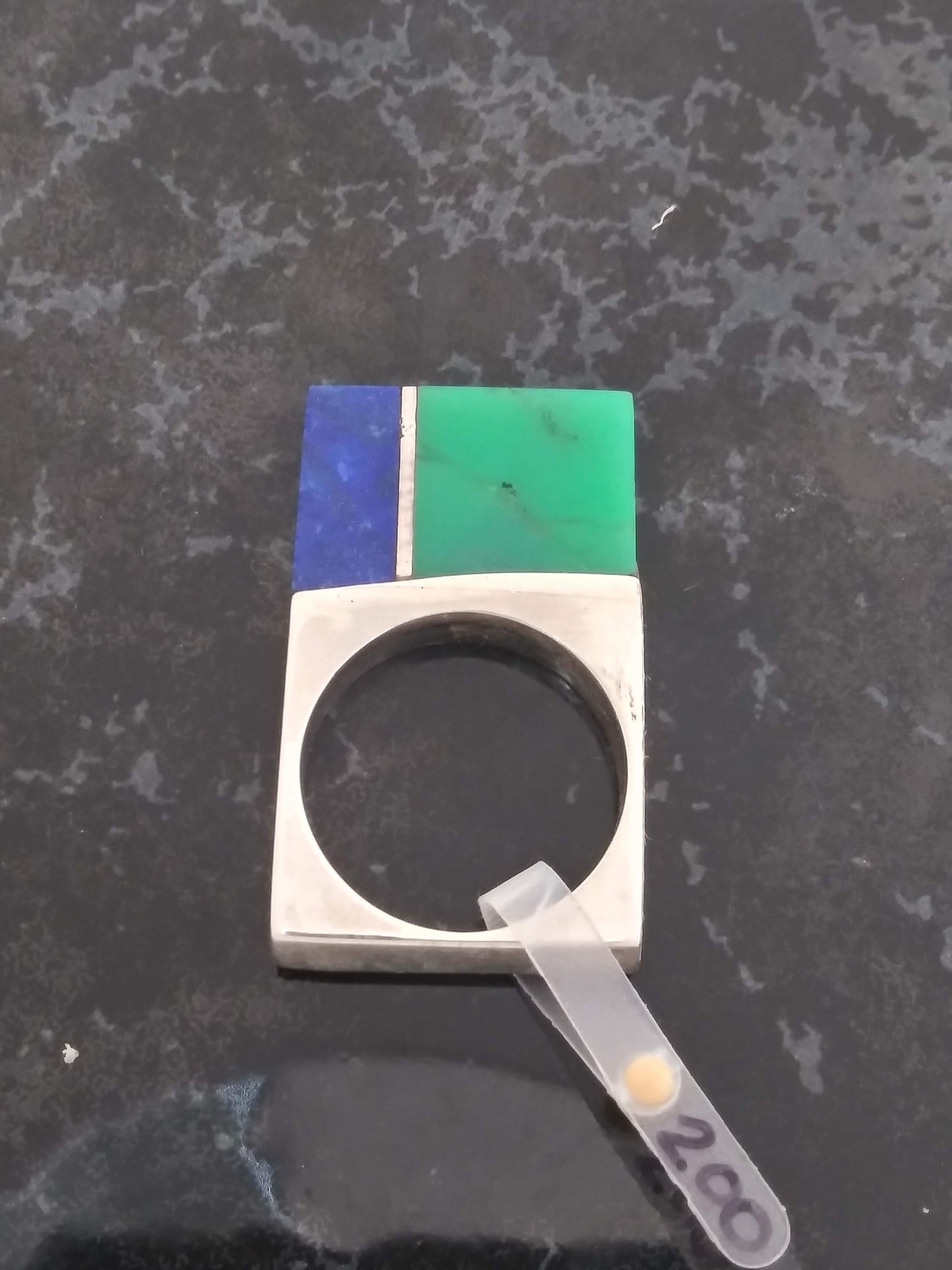 A silver ring with a large rectangular stone centerpiece. The stone is divided into two halves, one a deep blue lapis lazuli, and the other a vibrant green chrysoprase. A thin silver divider separates the two stones. The ring is sitting on a marble surface with a price tag that reads "200".
