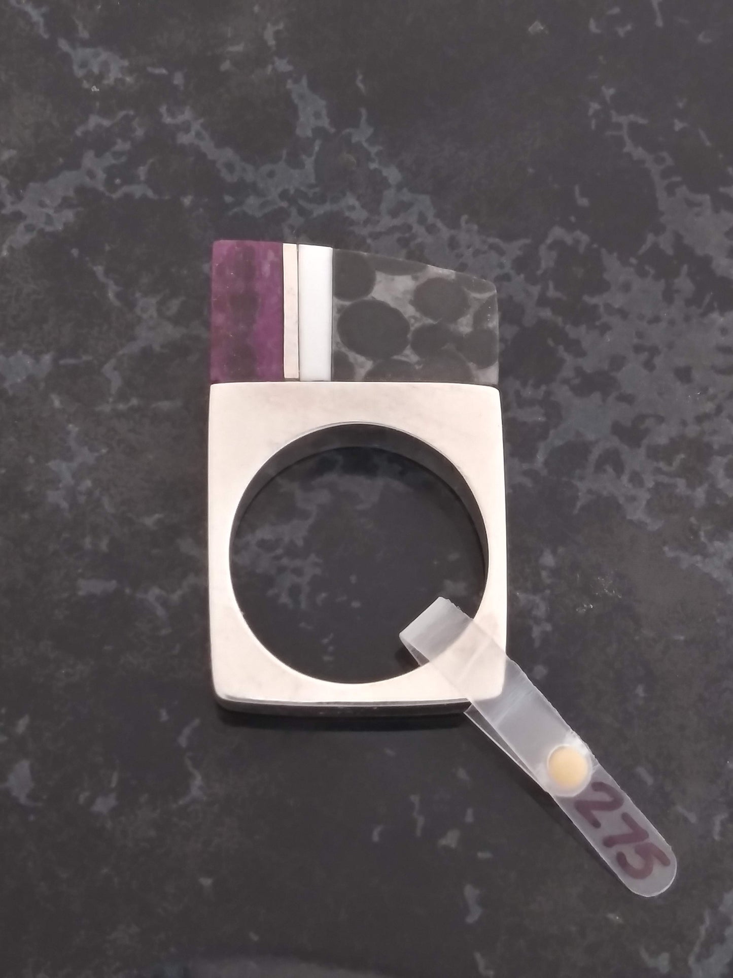 A silver ring with a square band and a rectangular stone setting featuring three vertical stripes of purple, white, and black stone. The ring is displayed on a black marble surface with a price tag reading "275".
