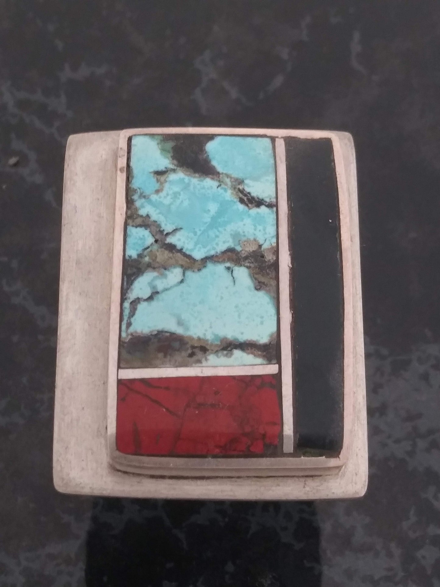 A square silver and turquoise inlay ring featuring red coral, black onyx, and turquoise stones arranged in a geometric pattern.