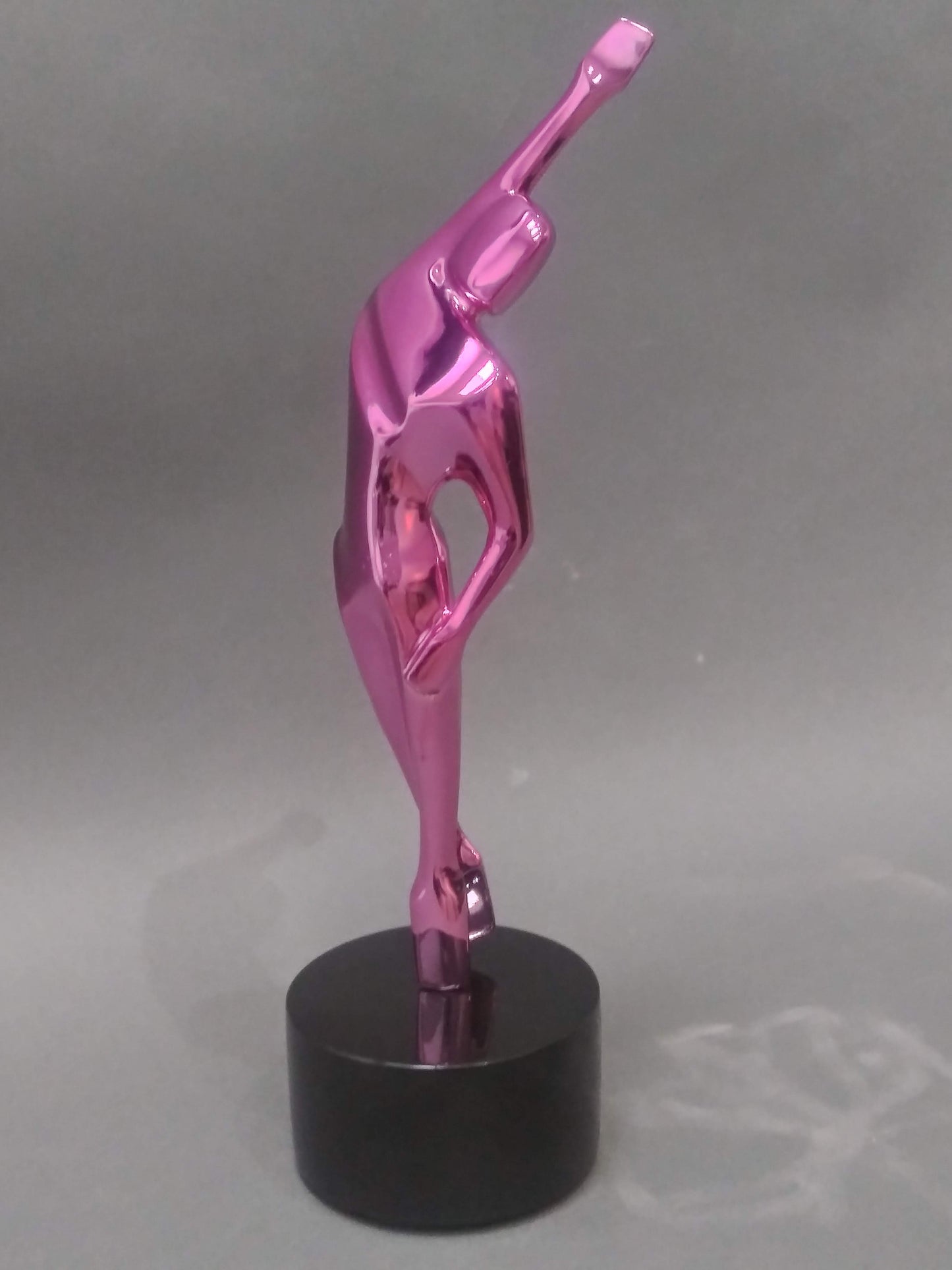 A metallic pink abstract sculpture of a person on a black base. The figure is standing on one leg with its arms raised above its head and its body arched backwards.