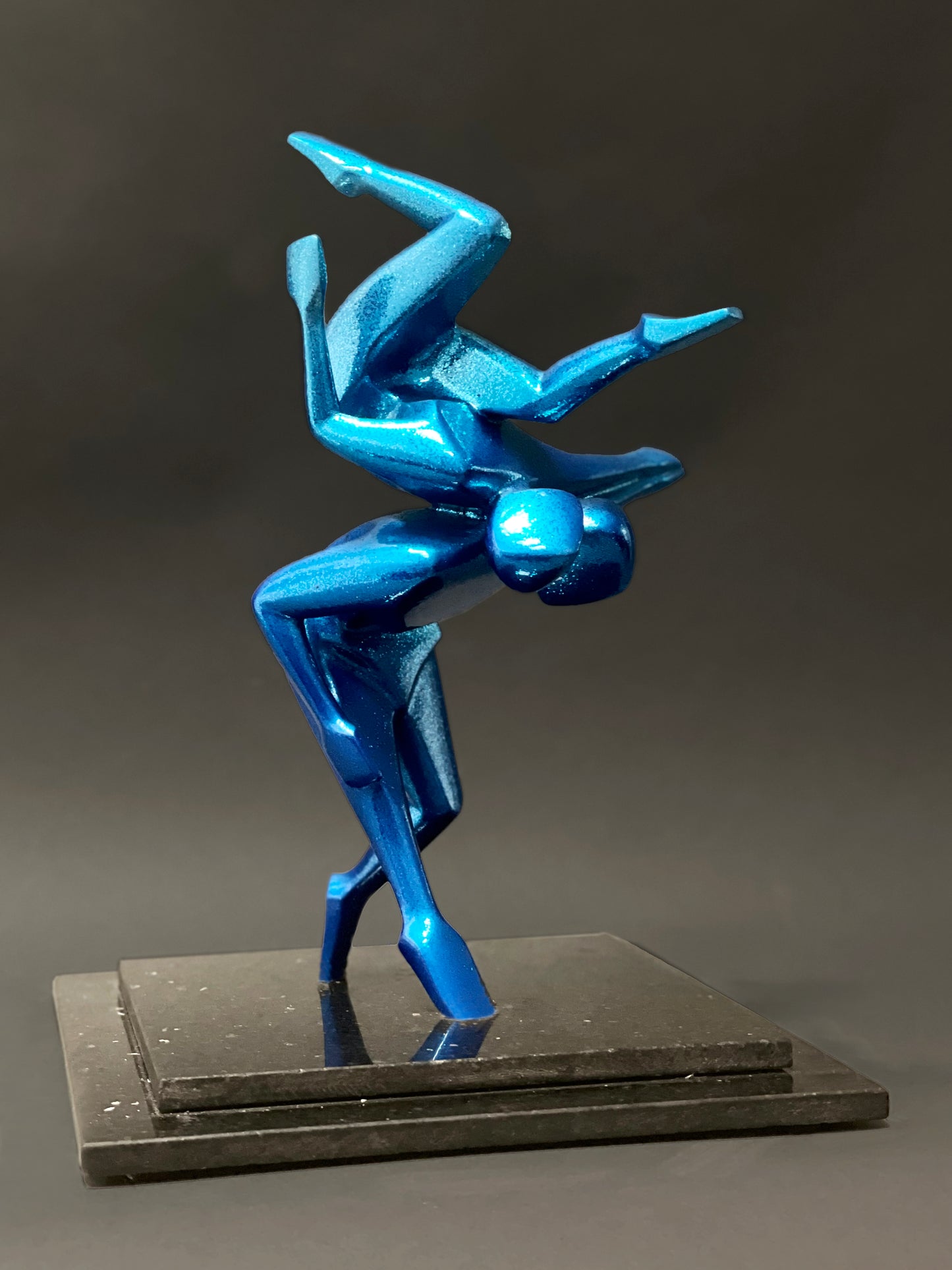 A metallic blue sculpture of two abstract human figures. One figure is balancing upside down on the shoulders of the other, who is crouched low. The sculpture is mounted on a black marble base.
