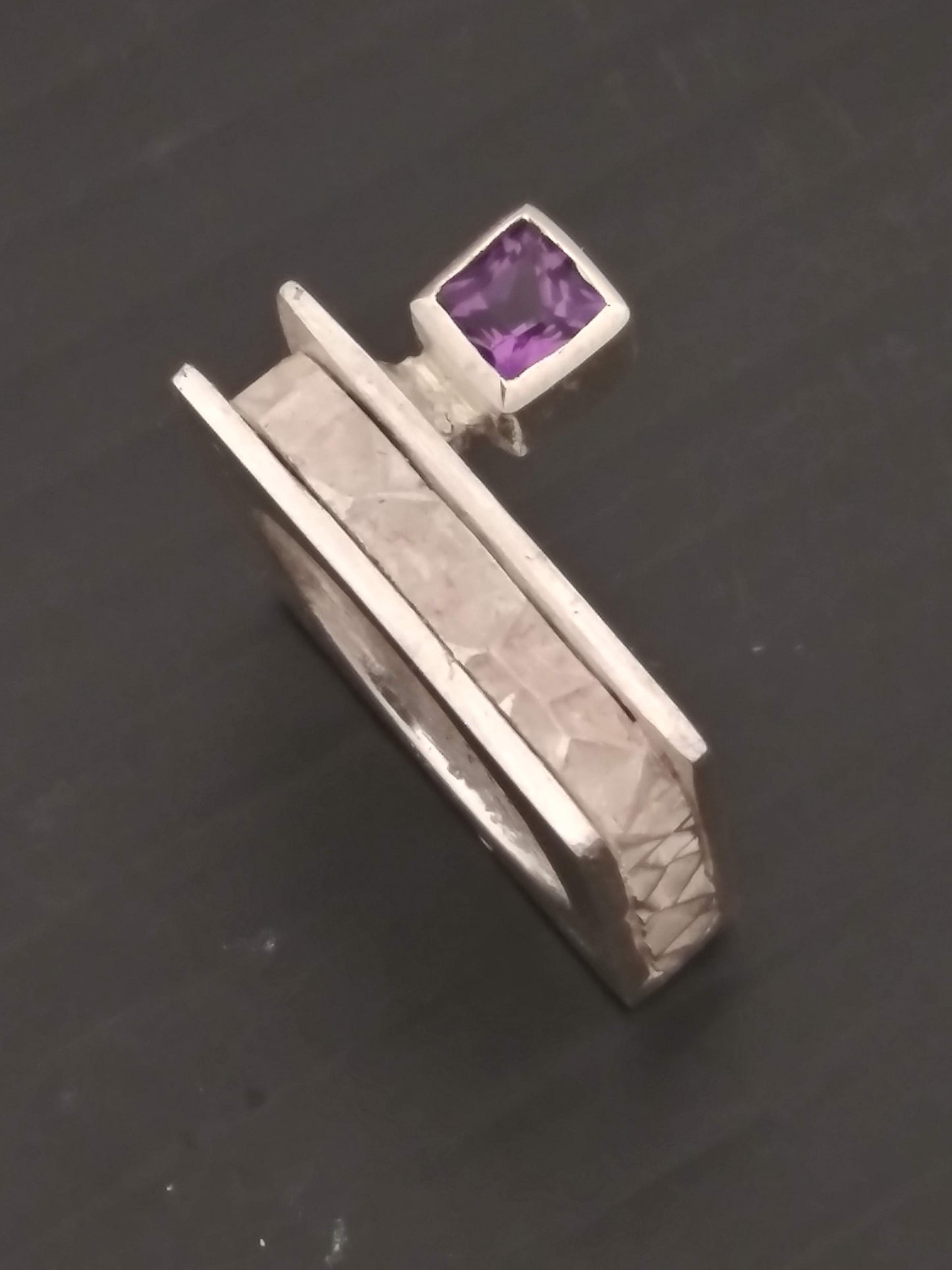 A silver ring with a square band featuring a textured pattern and a single square-cut amethyst gemstone.