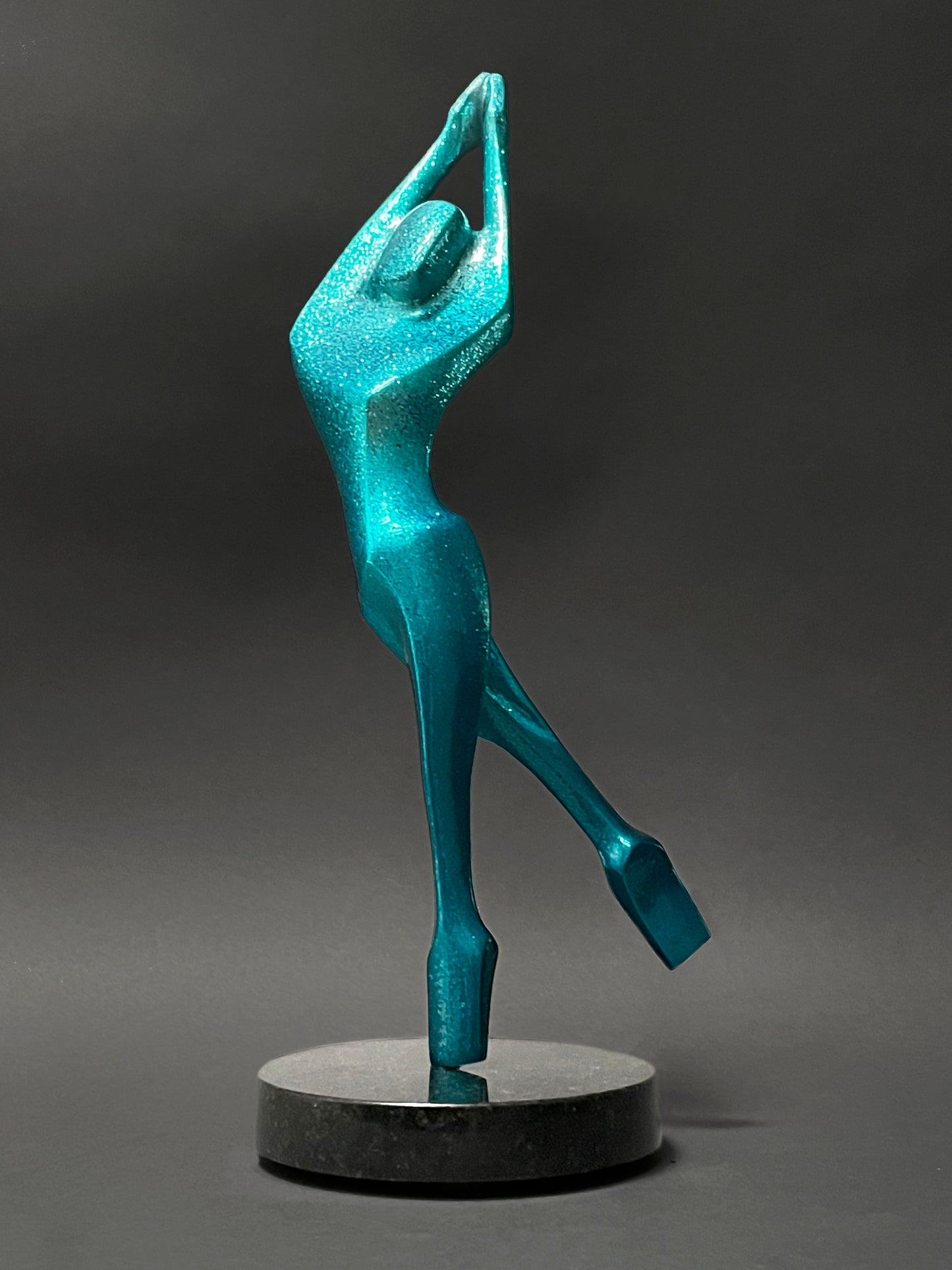 A bronze sculpture of a dancer with arms raised above their head, balancing on one pointed toe. The sculpture is a metallic teal color, mounted on a black base, and titled 'Turning Dancer' by Reno Carollo